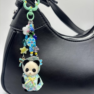 Op de afbeelding: Een tassenhanger met een bevlockt pandafiguurtje dat een lichtblauwe bloemige cape met capuchon en een geel lint draagt. De hanger hangt aan een mintgroene, bloemvormige sluiting en bevat een streng kleurrijke kralen, waaronder blauwe bloemen, een gele ster en een kleine blauwe strik.