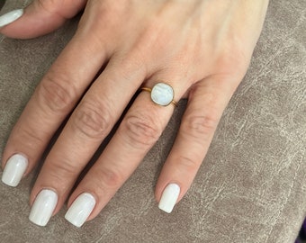 May include: A gold ring with a white moonstone gemstone. The ring is on a finger with white nail polish.