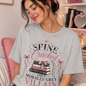 May include: A light gray t-shirt with the text "Spine Cracked for a Morally Grey Villain" in pink and black. The design includes a stack of books, bows, and a spine illustration. The shirt is worn with pink plaid pajamas.