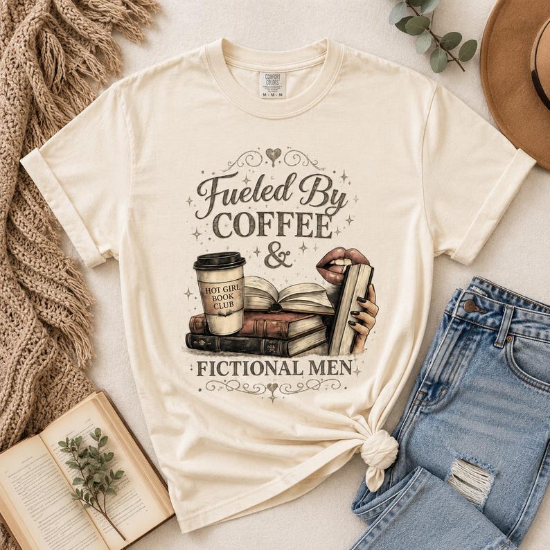 May include: Cream-colored t-shirt with the text "Fueled By Coffee & Fictional Men." The graphic includes a coffee cup, books, and lips. The shirt is paired with blue jeans and a hat.