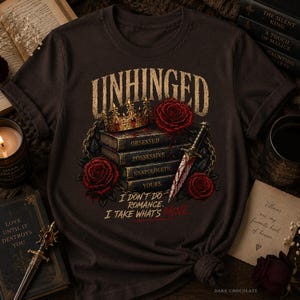 Puede incluir: Una camiseta de color marrón chocolate oscuro con un estampado gráfico de una corona dorada, rosas rojas, una daga y una pila de libros etiquetados como Obsessed, Possessive, Unapologetic y Yours. El texto dice Unhinged y I don't do romance. I take what's mine.