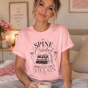 Pu&ograve; includere: T-shirt rosa chiaro con la scritta "Spine Cracked for a Morally Grey Villain" in un carattere decorativo. Il design include illustrazioni di libri e fiocchi. La persona tiene in mano una tazza con un cuore.