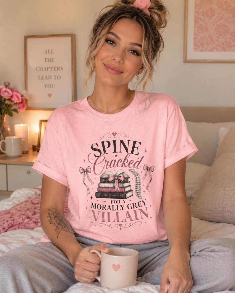 Pu&ograve; includere: T-shirt rosa chiaro con la scritta "Spine Cracked for a Morally Grey Villain" in un carattere decorativo. Il design include illustrazioni di libri e fiocchi. La persona tiene in mano una tazza con un cuore.