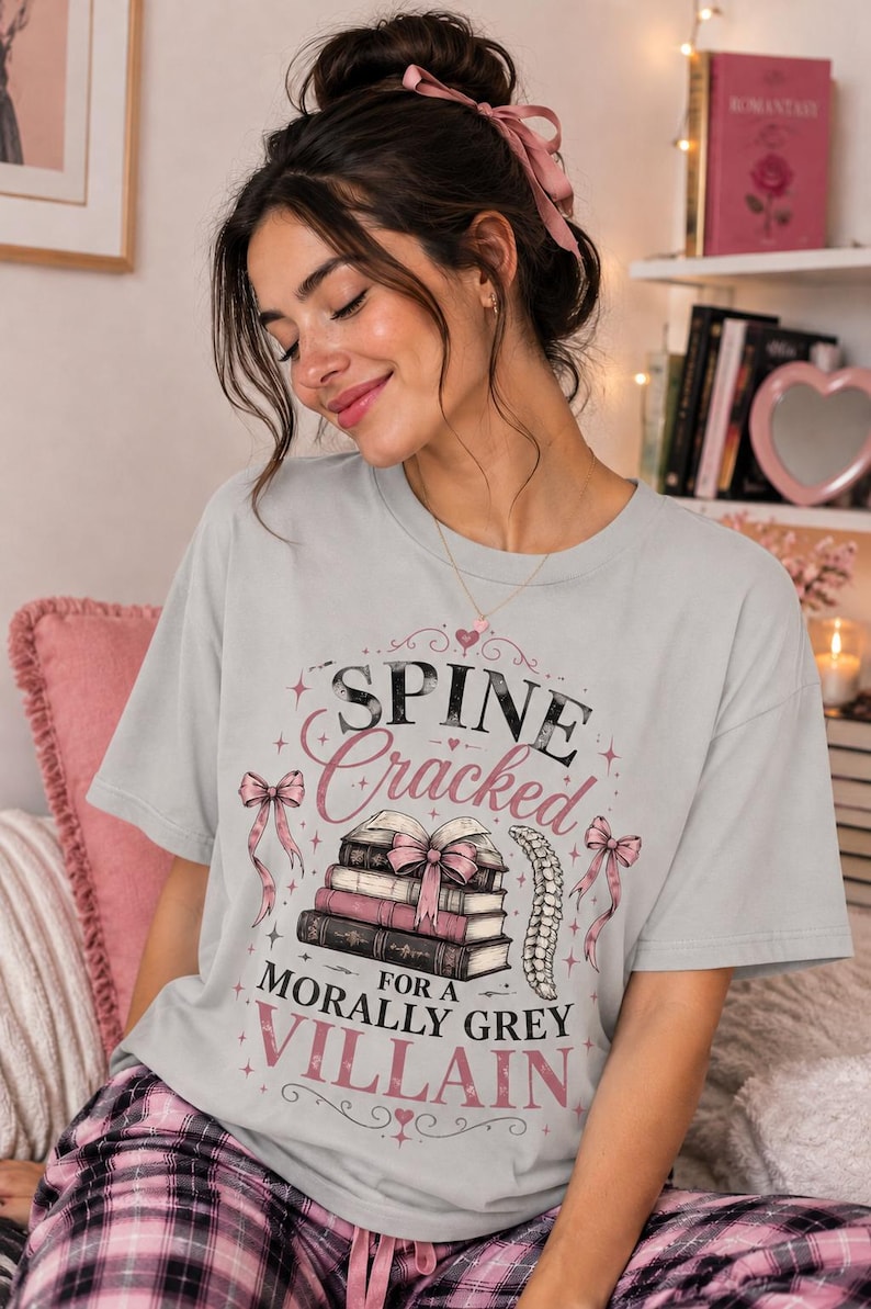 Pu&ograve; includere: T-shirt grigio chiaro con la scritta "Spine Cracked for a Morally Grey Villain" in rosa e nero. Il design include una pila di libri, fiocchi e un'illustrazione della colonna vertebrale. La maglietta &egrave; indossata con un pigiama a quadri rosa.