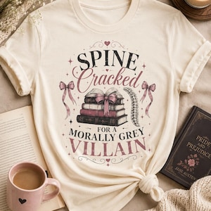 Pu&ograve; includere: T-shirt color crema con la scritta "Spine Cracked for a Morally Grey Villain". La grafica include una pila di libri legati con un fiocco rosa, una spina dorsale ed elementi decorativi. Sono visibili anche un libro e una tazza.
