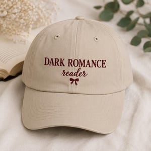 May include: A beige baseball cap featuring the embroidered text DARK ROMANCE reader in dark red, with a small dark red bow icon centered below the text. The cap has a structured crown, a curved brim, and stitched eyelets on the top panels.