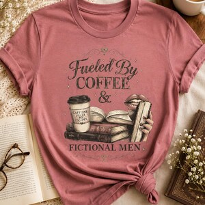 May include: A dusty rose t-shirt with the text "Fueled By Coffee & Fictional Men." The graphic includes a coffee cup, books, and lips. The shirt is knotted at the bottom and is surrounded by books and a cup of coffee.