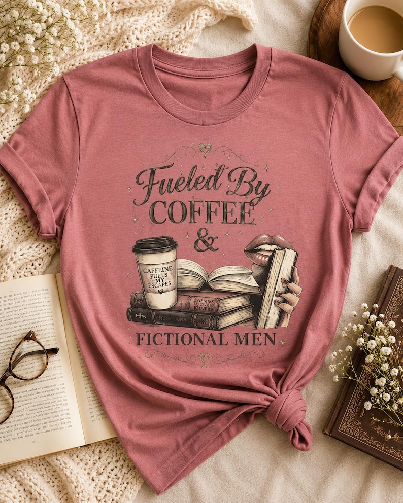 May include: A dusty rose t-shirt with the text "Fueled By Coffee & Fictional Men." The graphic includes a coffee cup, books, and lips. The shirt is knotted at the bottom and is surrounded by books and a cup of coffee.