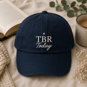 May include: A navy blue baseball cap featuring white embroidered text that reads TBR Today, centered above a small four-pointed star icon. The cap has a structured crown, a curved brim, and stitched eyelets on the top panels for ventilation.