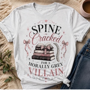 Pu&ograve; includere: T-shirt bianca con la scritta "Spine Cracked for a Morally Grey Villain" in nero e rosa. Il design include una pila di libri legati con un fiocco, un'illustrazione della colonna vertebrale ed elementi decorativi.