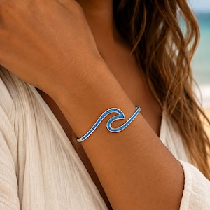 May include: A silver-toned open cuff bracelet featuring a stylized wave design. The center of the wave shape is inlaid with vibrant blue crushed material that creates a textured, sparkling appearance against the polished metallic frame.