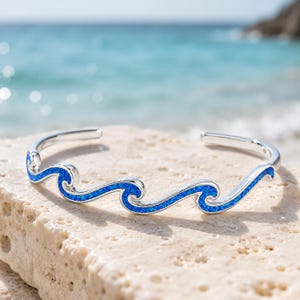 May include: A silver cuff bracelet featuring a continuous wave design. The wave pattern is inlaid with vibrant blue crushed material that sparkles, creating a distinct contrast against the polished silver metal of the open-ended band.