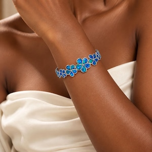 Puede incluir: Una pulsera de tono plateado que presenta una serie de eslabones conectados en forma de flor. Cada eslabón floral está incrustado con piedras iridiscentes de color azul y turquesa similares al ópalo, creando un patrón floral continuo.