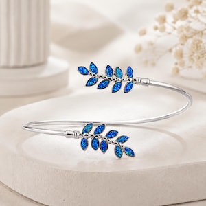 May include: A silver-toned open cuff bracelet featuring two symmetrical leaf-shaped motifs at each end. Each leaf is inlaid with vibrant blue iridescent stones that shimmer with a speckled pattern, creating a delicate botanical design.