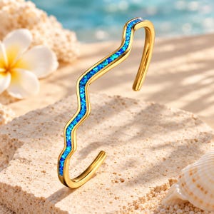 Puede incluir: Una pulsera abierta de tono dorado con un diseño ondulado continuo. El centro de la onda está incrustado con un material texturizado de color azul vibrante que imita la apariencia de piedra triturada o purpurina, creando un contraste llamativo.