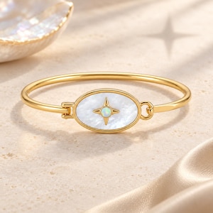 Puede incluir: Una pulsera rígida de tono dorado con una pieza central ovalada con incrustaciones de nácar blanco. En el centro del óvalo hay un motivo de estrella de cuatro puntas hecho de metal dorado, engastado con una pequeña gema de ópalo redonda.