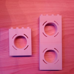 May include: Two white, 3D-printed, rectangular blocks with circular cutouts and curved lines. One block is a single unit, while the other is a stack of two. The blocks have small, raised studs on top, resembling building blocks. They rest on a light brown wooden surface.