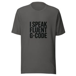 May include: A dark gray t-shirt with the text "I SPEAK FLUENT G-CODE" printed in black. The shirt has short sleeves and a crew neck. The text is centered on the front of the shirt.