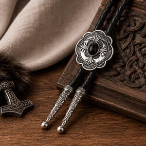 May include: A bolo tie featuring a round, scalloped silver-tone metal centerpiece with an oval black stone inlay. The tie consists of a braided black leather cord finished with two matching silver-tone metal tips with intricate engraved patterns.