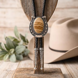 May include: A bolo tie featuring a central oval-shaped brown and tan banded stone set within a silver-toned horseshoe frame. The cord is made of braided black leather and includes two matching silver-toned metal tips with engraved detailing.