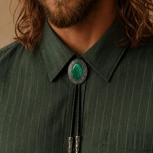 May include: A bolo tie featuring an oval-shaped green malachite stone set in an ornate, silver-toned metal frame with intricate scrollwork. The tie includes a black braided cord finished with two matching silver-toned metal tips with engraved details.