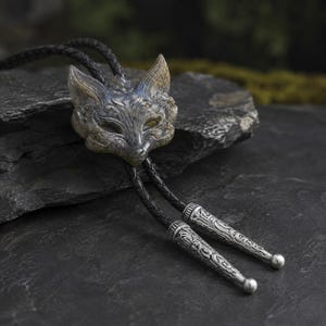 May include: A bolo tie featuring a carved stone fox head pendant with intricate swirling patterns. The pendant is attached to a black braided leather cord that terminates in two pointed, engraved silver-toned metal tips with rounded ends.