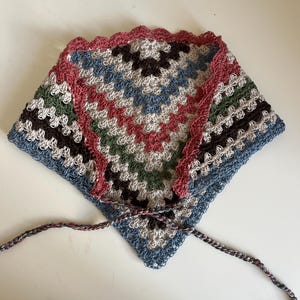 May include: A triangular crochet bandana featuring a multi-colored granny square pattern in shades of dusty rose, sage green, dark brown, light gray, and denim blue. The edges are finished with a scalloped rose border and two long braided ties.