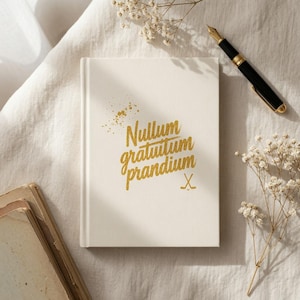 May include: A cream-colored hardcover notebook featuring the gold-colored text Nullum gratuitum prandium and a small gold hockey stick icon. A black fountain pen with gold trim rests nearby, accompanied by sprigs of white baby's breath flowers.