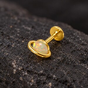 May include: A gold-tone labret stud earring featuring a planet-shaped design. The center is a round, iridescent white opal cabochon surrounded by a thin gold-tone ring, attached to a straight post with a flat circular back disc.