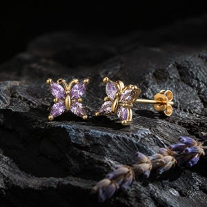 May include: A pair of gold stud earrings shaped like butterflies, each featuring four marquise-cut light purple stones forming the wings and a small gold center. The earrings have a post back and are displayed with a sprig of dried lavender.