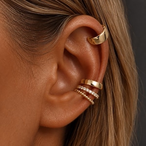 May include: A set of four gold-toned ear cuffs worn on the cartilage and conch. The top cuff is a smooth, polished band. The lower three cuffs include a plain polished band, a band encrusted with small clear stones, and a delicate beaded band.