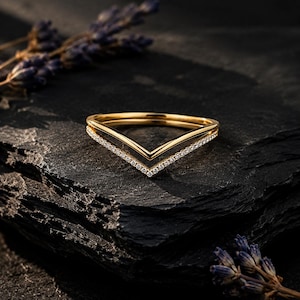 May include: A gold V-shaped ring featuring a double band design. One band is a smooth, polished gold wire, while the second band is set with a row of small, clear, sparkling stones that follow the chevron silhouette of the ring.