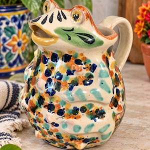 May include: A ceramic pitcher shaped like a frog with a cream-colored base. The surface is decorated with hand-painted spots in shades of blue, orange, and teal. It features a wide mouth, protruding eyes, and a curved handle on the right side.