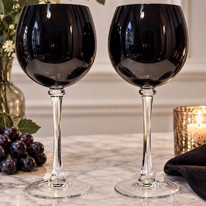 May include: Two wine glasses with large, rounded, dark charcoal-colored bowls and clear, slender stems that transition into circular, transparent glass bases. The glasses feature a smooth, reflective surface and a classic, tall silhouette.