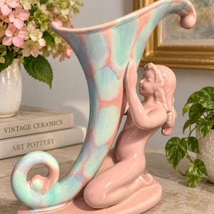 May include: A ceramic cornucopia vase featuring a pastel pink, blue, and white mottled glaze. The vase is supported by a kneeling figure with a smooth, solid pink finish. The cornucopia has a curved, horn-like shape with a flared opening and a curled base.