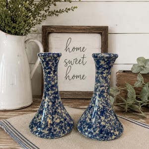 May include: A pair of ceramic candle holders featuring a speckled blue and cream pattern. Each holder has a flared base that tapers upward to a circular top opening. The surface of the ceramic is covered in a dense, mottled blue and white design.