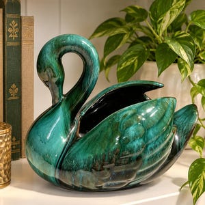May include: A ceramic swan planter featuring a glossy, deep emerald green glaze with dark black undertones and a smooth, curved neck. The swan has a hollow center for holding items, with detailed molded wing textures and a polished, reflective finish.