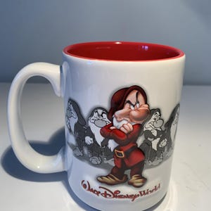 May include: A white ceramic mug with a red interior and handle. The exterior features a colorful illustration of Grumpy from Snow White and the Seven Dwarfs, surrounded by grayscale sketches of the character. The text Walt Disney World is printed below.