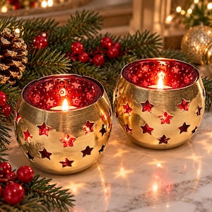 May include: Two spherical candle holders featuring a metallic gold exterior with cut-out star shapes and a deep red interior. Each holder contains a lit tea light candle, casting a warm glow through the star-shaped openings onto the surface.