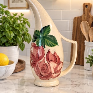 May include: A tall, cream-colored ceramic pitcher with a curved spout and a handle. The body of the pitcher features a hand-painted design of deep red roses with green leaves. The pitcher has a pedestal base and a smooth, glossy finish throughout.