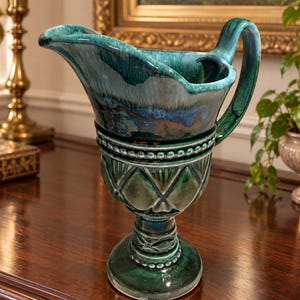 May include: A ceramic pitcher featuring a deep teal and forest green glaze with a glossy finish. The vessel has a wide spout, a curved handle, and a body decorated with embossed diamond patterns and beaded detailing around the neck and base.