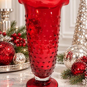 May include: A tall, vibrant red glass vase featuring a textured hobnail pattern across its surface. The vase has a wide, ruffled, scalloped rim and sits on a circular pedestal base. The glass is translucent, allowing light to pass through.
