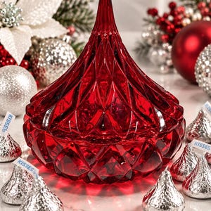 May include: A deep red, faceted glass container shaped like a Hershey's Kiss, featuring a pointed top and a diamond-patterned surface. The container is surrounded by several silver-wrapped chocolate candies, each with a small white paper tag.