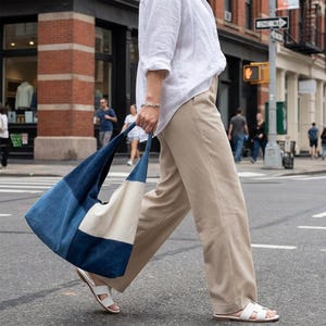 Könnte beinhalten: Eine große Tragetasche mit einem Patchwork-Design in Jeansblau und Creme. Die Tasche wird von einer Person getragen, die eine Stadtstraße überquert. Das Design der Tasche zeigt einen großen jeansblauen Abschnitt und einen cremefarbenen Abschnitt.