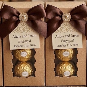 Peut inclure: Deux boîtes rectangulaires en papier kraft contenant chacune deux chocolats enveloppés dans du papier doré. Chaque boîte présente un nœud en satin marron avec une décoration en strass et une étiquette Alicia and Jason Engaged October 11th 2026.