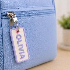 May include: A light blue backpack with a silver zipper and a mesh side pocket. A rectangular tag with the name "OLIVIA" in purple letters hangs from the zipper. A small potted plant is in the background.
