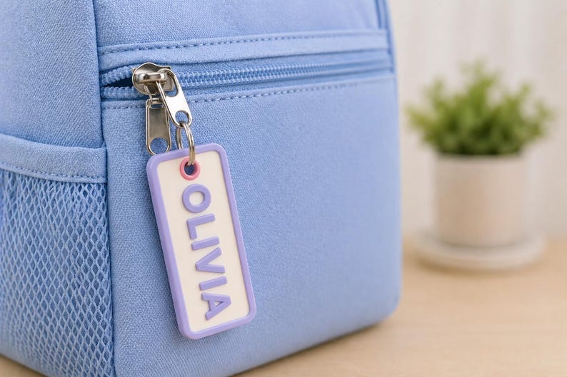 May include: A light blue backpack with a silver zipper and a mesh side pocket. A rectangular tag with the name "OLIVIA" in purple letters hangs from the zipper. A small potted plant is in the background.