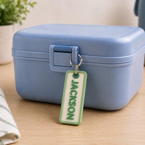 May include: A light blue lunch box with a matching latch and a green and white tag that reads "JACKSON". The lunch box is rectangular and sits on a light wood surface. A small plant in a white pot is in the background.