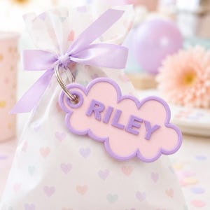 Pu&ograve; includere: Un sacchetto regalo con un motivo a cuori bianchi e pastello, legato con un nastro lilla. Un'etichetta a forma di nuvola con il nome "RILEY" in lettere lilla &egrave; attaccata. Lo sfondo include un fiore rosa e un palloncino viola.