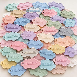 May include: A collection of pastel-colored cloud-shaped name tags. Each tag features a different name in a sans-serif font. The tags are in various colors, including pink, blue, yellow, green, and purple. They have a small hole at the top for attaching to items.